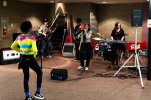 Four teenagers playing lead guitar, bass, keyboard, and singing lead vocals while patrons listen