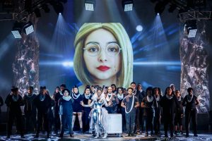 A woman with blond hair and glasses projected on a screen behind a group of performers mostly wearing black. A woman in a metallic bodysuit with a ruff stand front and center