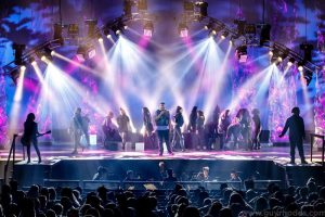 A filled stage with streams of blue and purple light. A packed audience enrapt. 